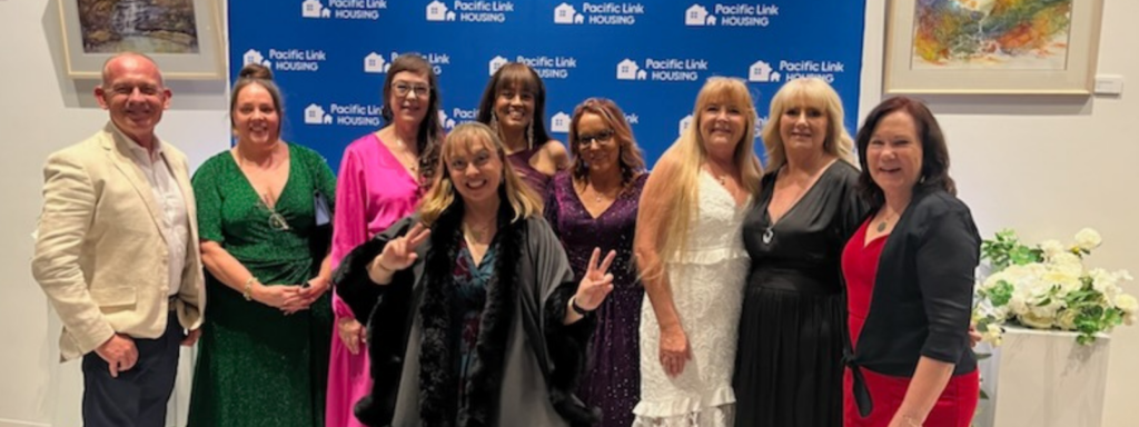 Nine smiling members of the Coastlink team pose in front of a branded blue-and-white photo wall